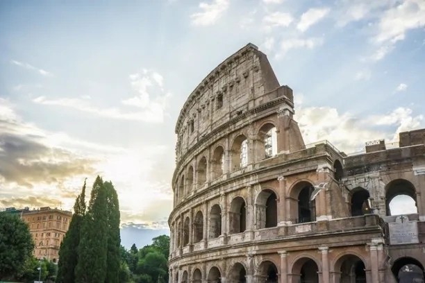 Colosseum with arches under a cloudy sky and trees in the background during sunset.