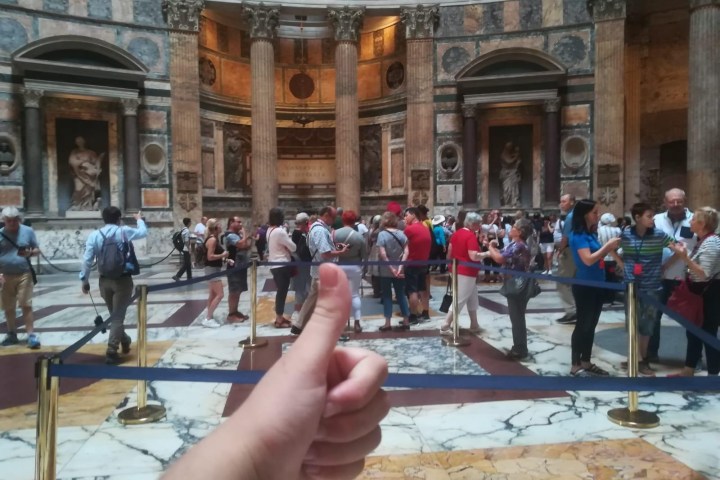 Thumbs-up hand gesture in front of a crowd inside a historic building with columns and statues.